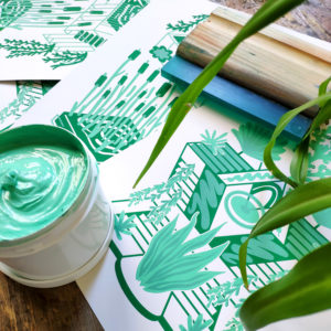 affiches sérigraphiées en vert disposées sur une table en bois accompagné d'une racle de sérigraphie, d'encre à l'eau et d'une plante.